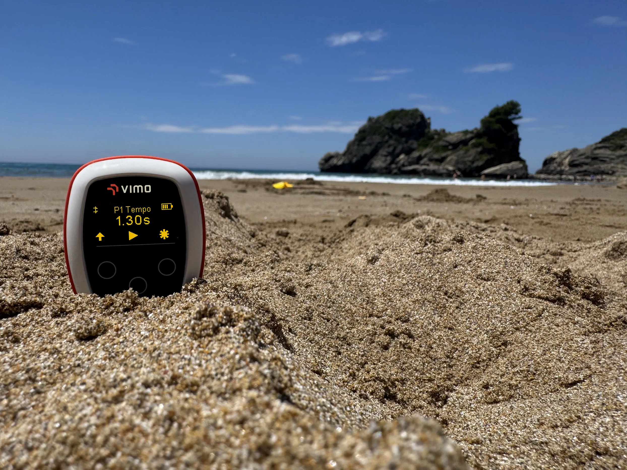 VimoSwim tempo trainer in the sand on a beach, OLED showing P1 Tempo 1.30s, ocean and headland behind
