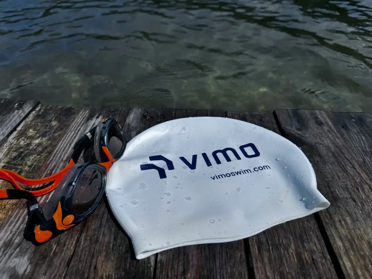 A Vimo Swim cap and goggles drying on a wooden jetty above open water