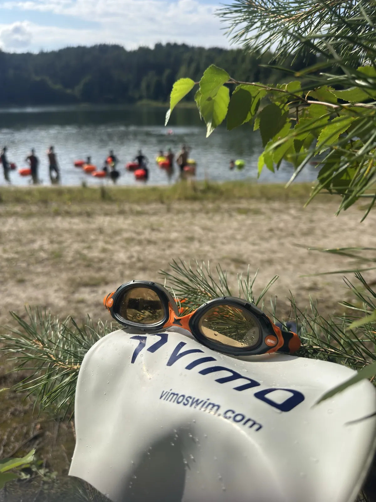 Vimo dry bag and goggles on the shore, with swimmers gathering by the lake behind